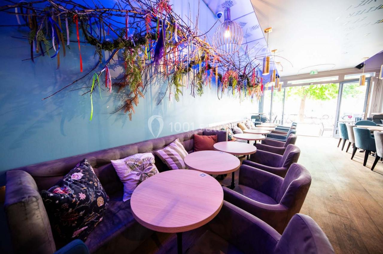 Salle de café lumineuse avec des tables rondes en bois, des fauteuils rembourrés et une décoration florale suspendue.