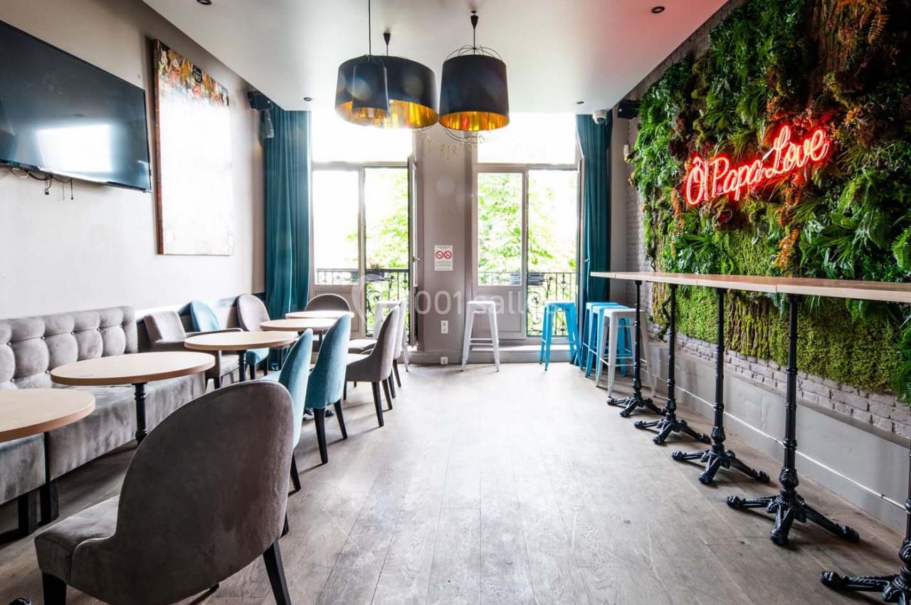 Salle lumineuse avec tables, chaises, mur végétal, enseigne néon rouge et grandes fenêtres donnant sur l'extérieur.