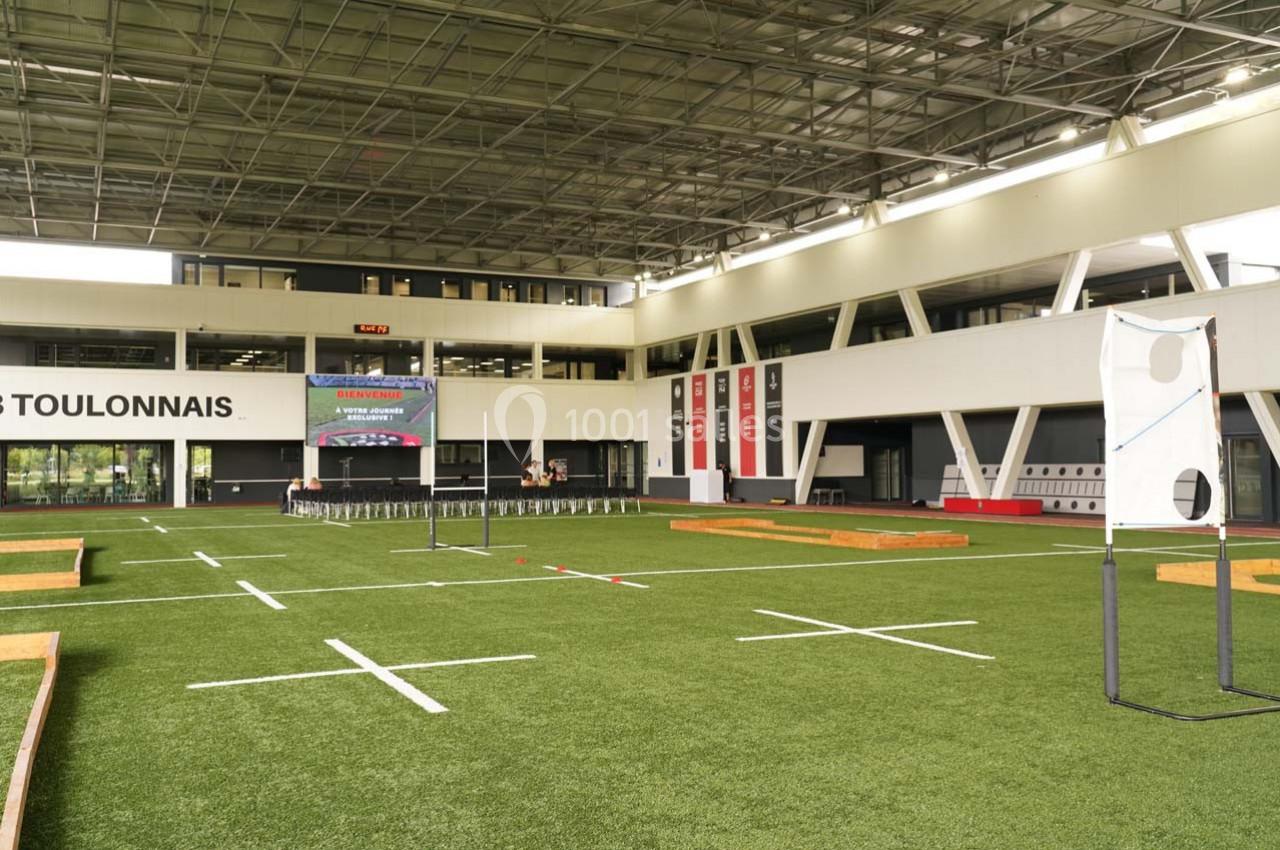 Terrain intérieur avec pelouse synthétique, zones délimitées, structures d'entraînement et gradins en arrière-plan.