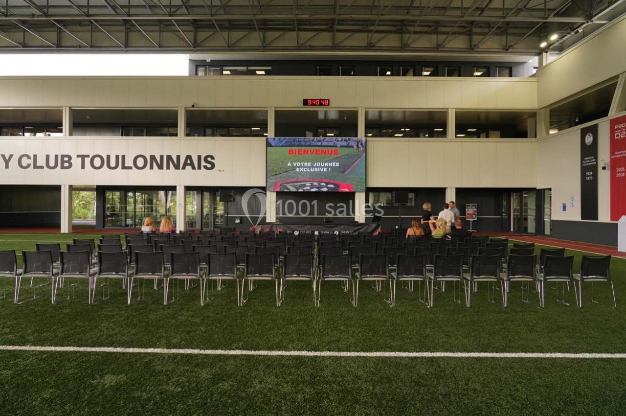 Salle de conférence en plein air avec écran géant, chaises alignées et bâtiment moderne en arrière-plan.