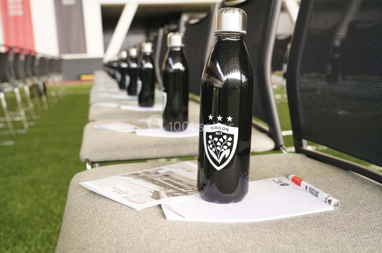 Bouteille noire posée sur une chaise avec un carnet, un stylo et des sièges alignés en arrière-plan.