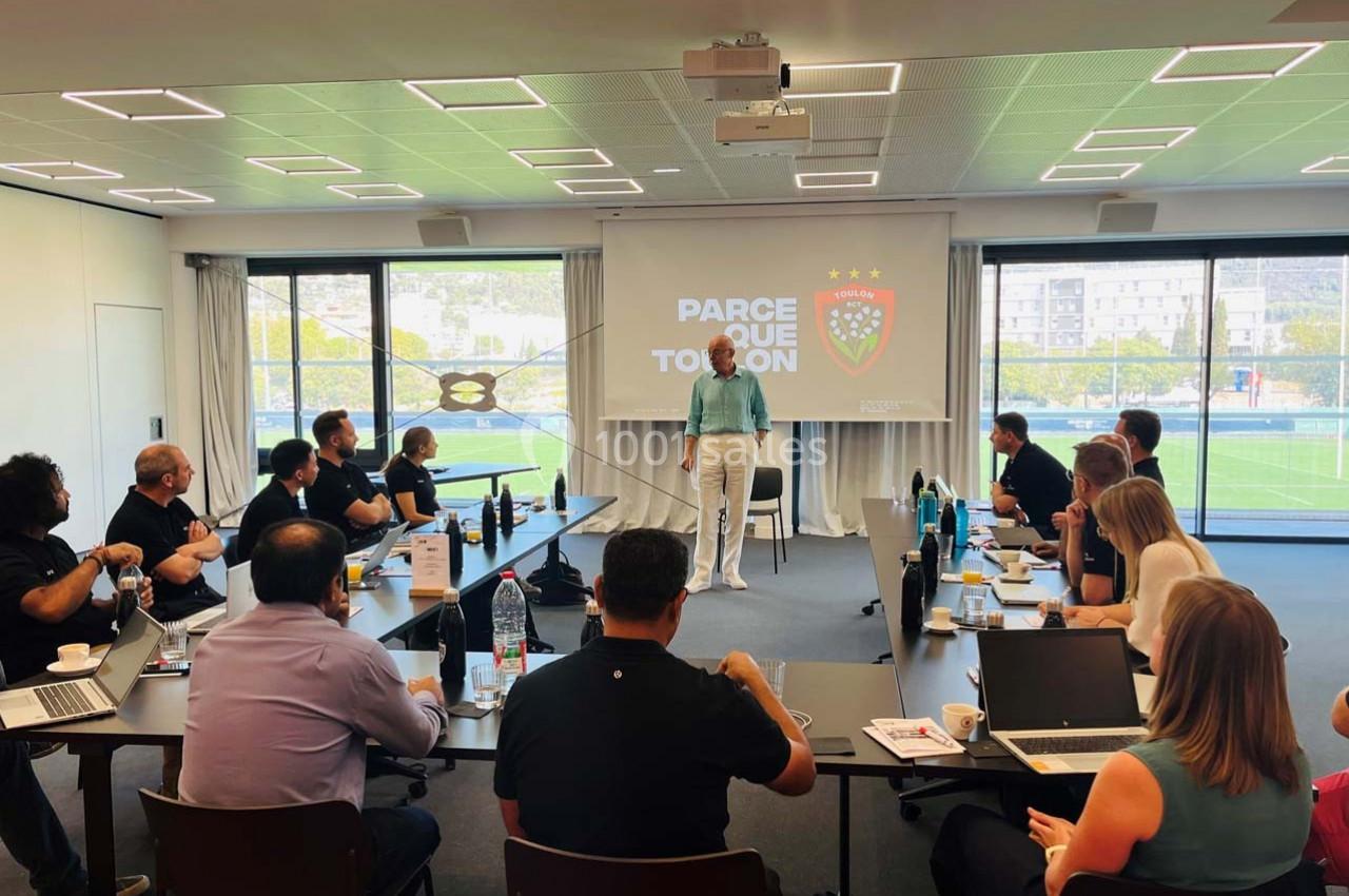 Une personne debout présente devant un groupe assis dans une salle de réunion avec écran et grandes baies vitrées.