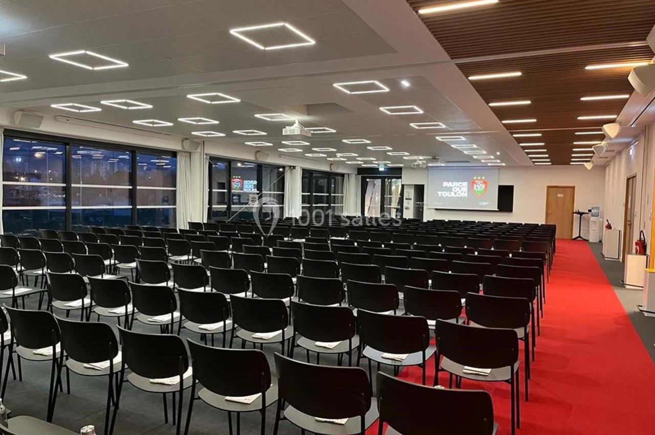 Salle de conférence moderne avec rangées de chaises noires, écran de projection et éclairage au plafond.