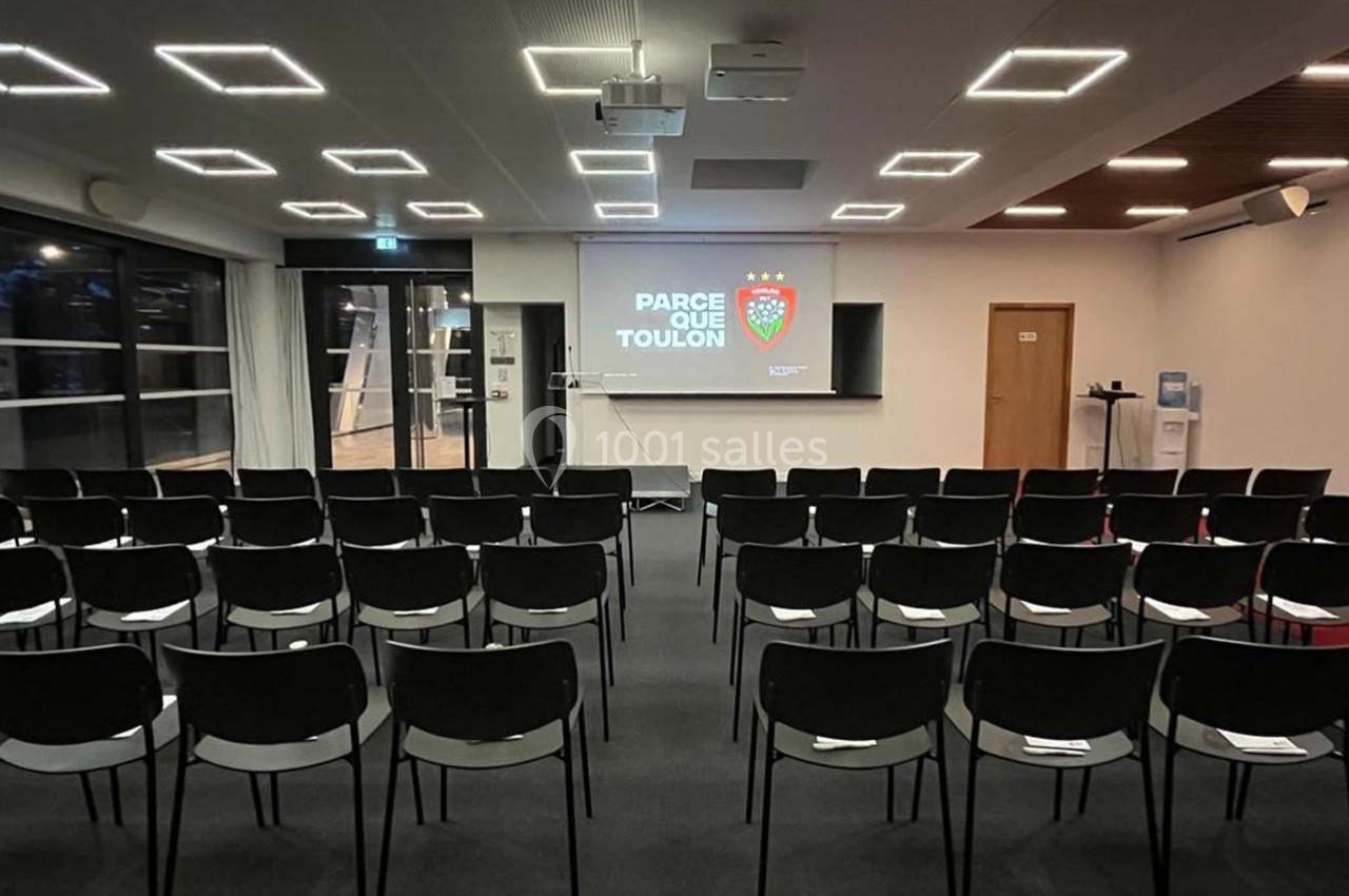 Salle de conférence vide avec des rangées de chaises noires, un écran affichant ’Parce que Toulon’ et un projecteur allumé.