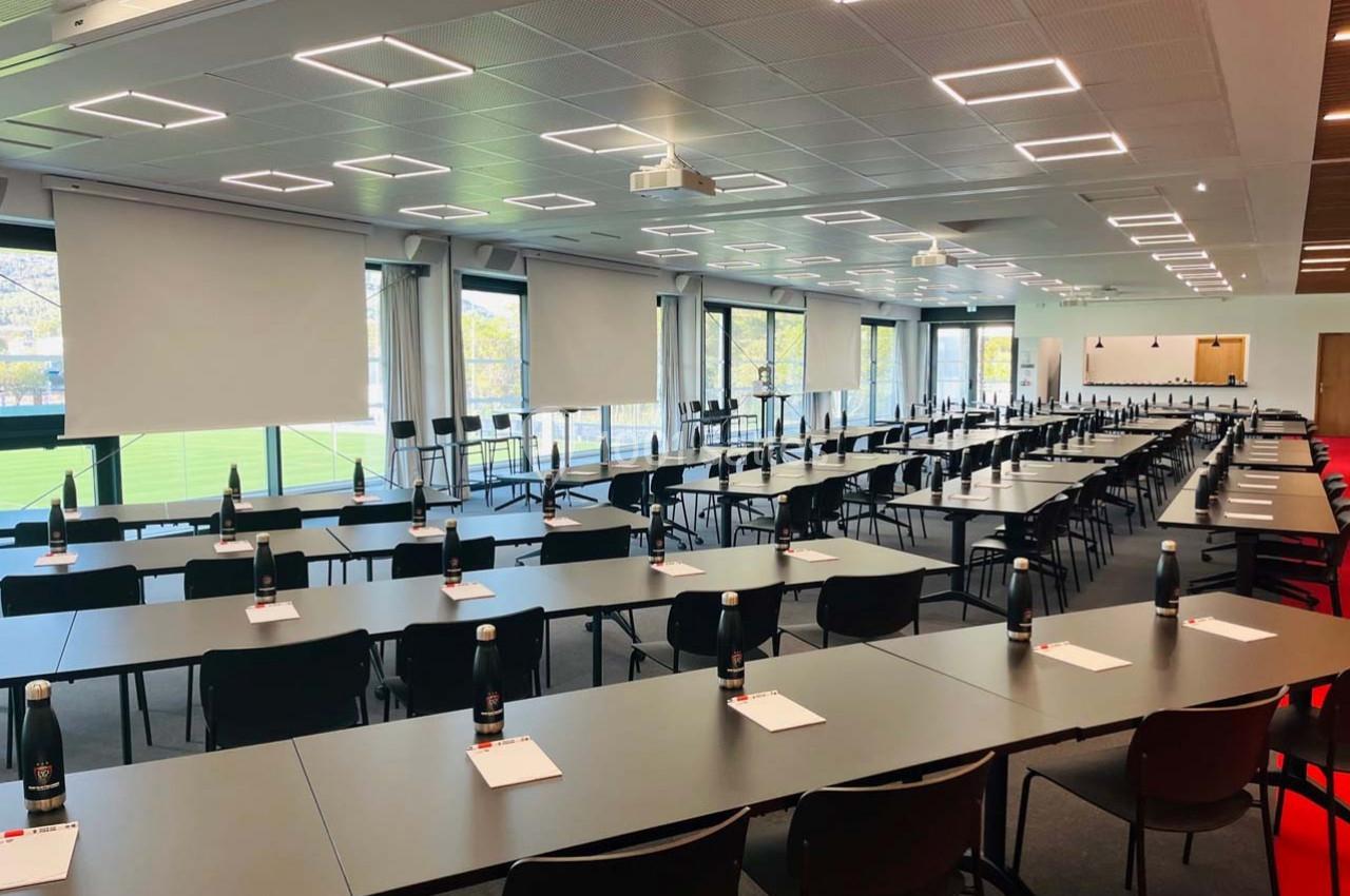 Salle de conférence moderne avec des rangées de tables et chaises, blocs-notes et bouteilles d'eau disposés.