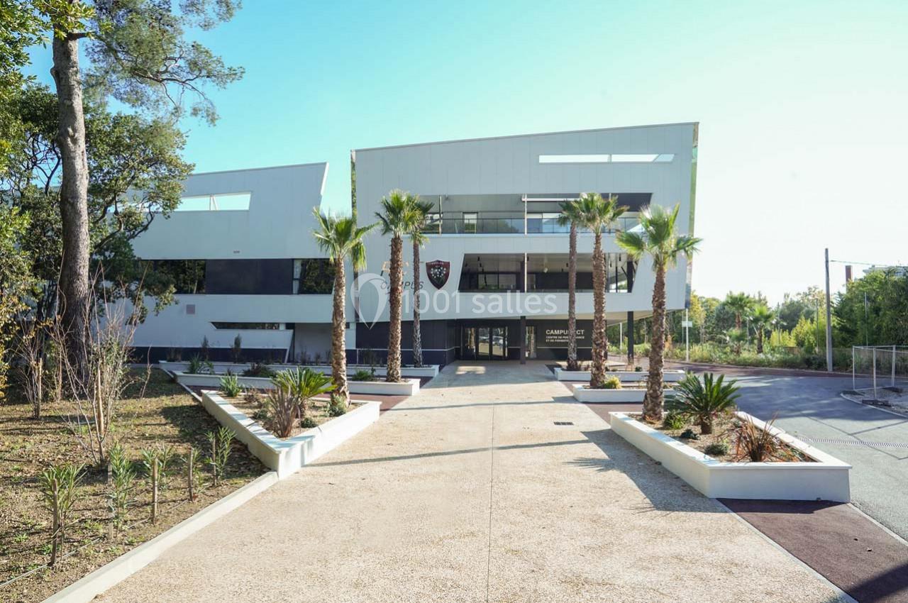 Bâtiment moderne entouré de palmiers, avec une allée centrale bordée de plantes et un ciel dégagé en arrière-plan.