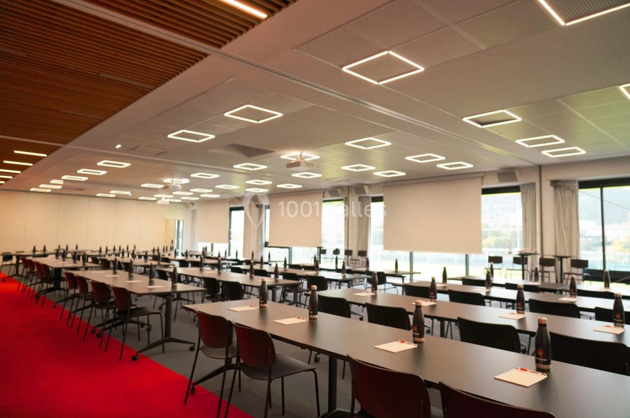 Salle de conférence moderne avec des rangées de tables et chaises, éclairage géométrique et grandes fenêtres.