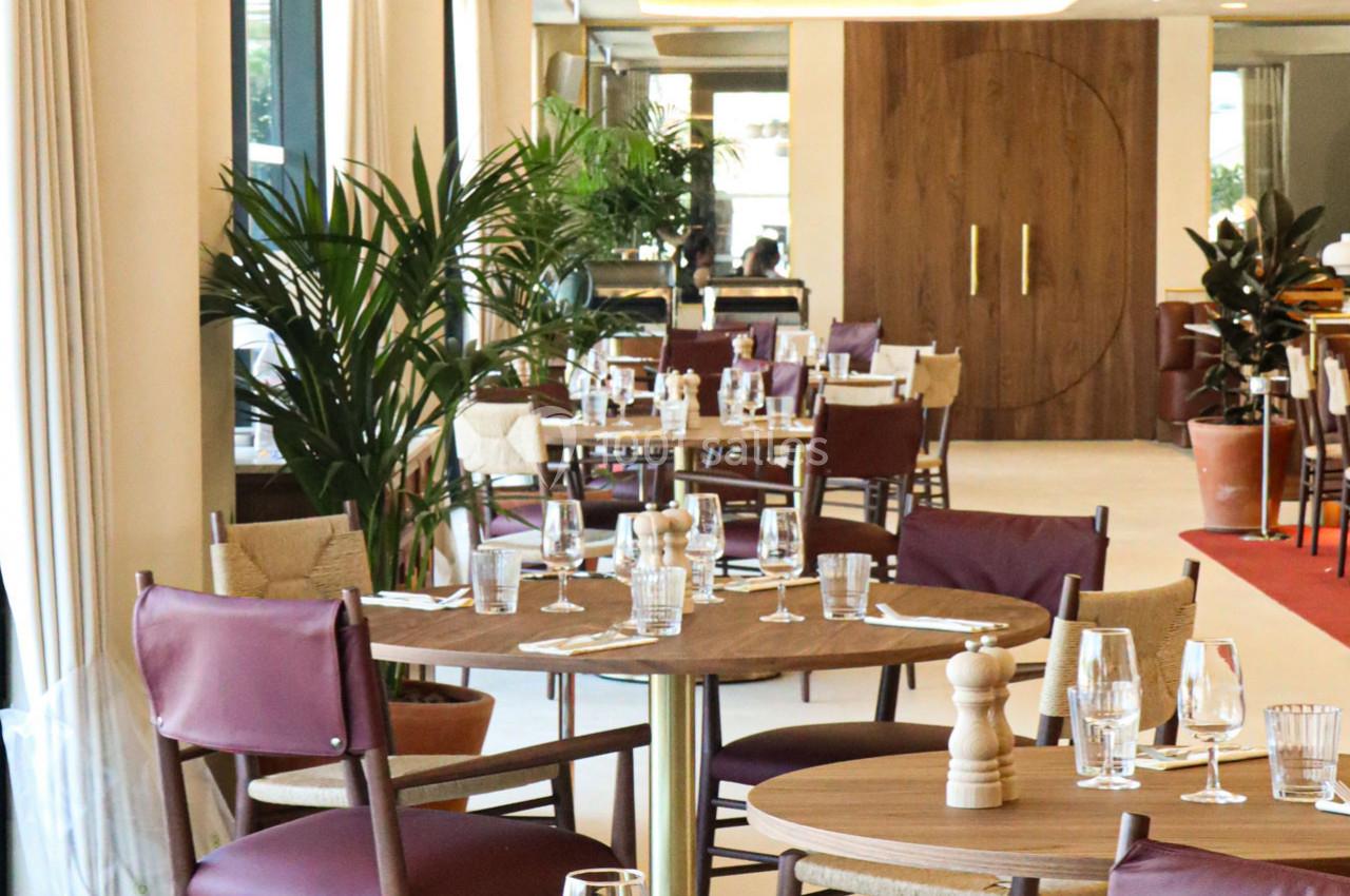Salle de restaurant lumineuse avec tables en bois dressées, chaises bordeaux et plantes décoratives.