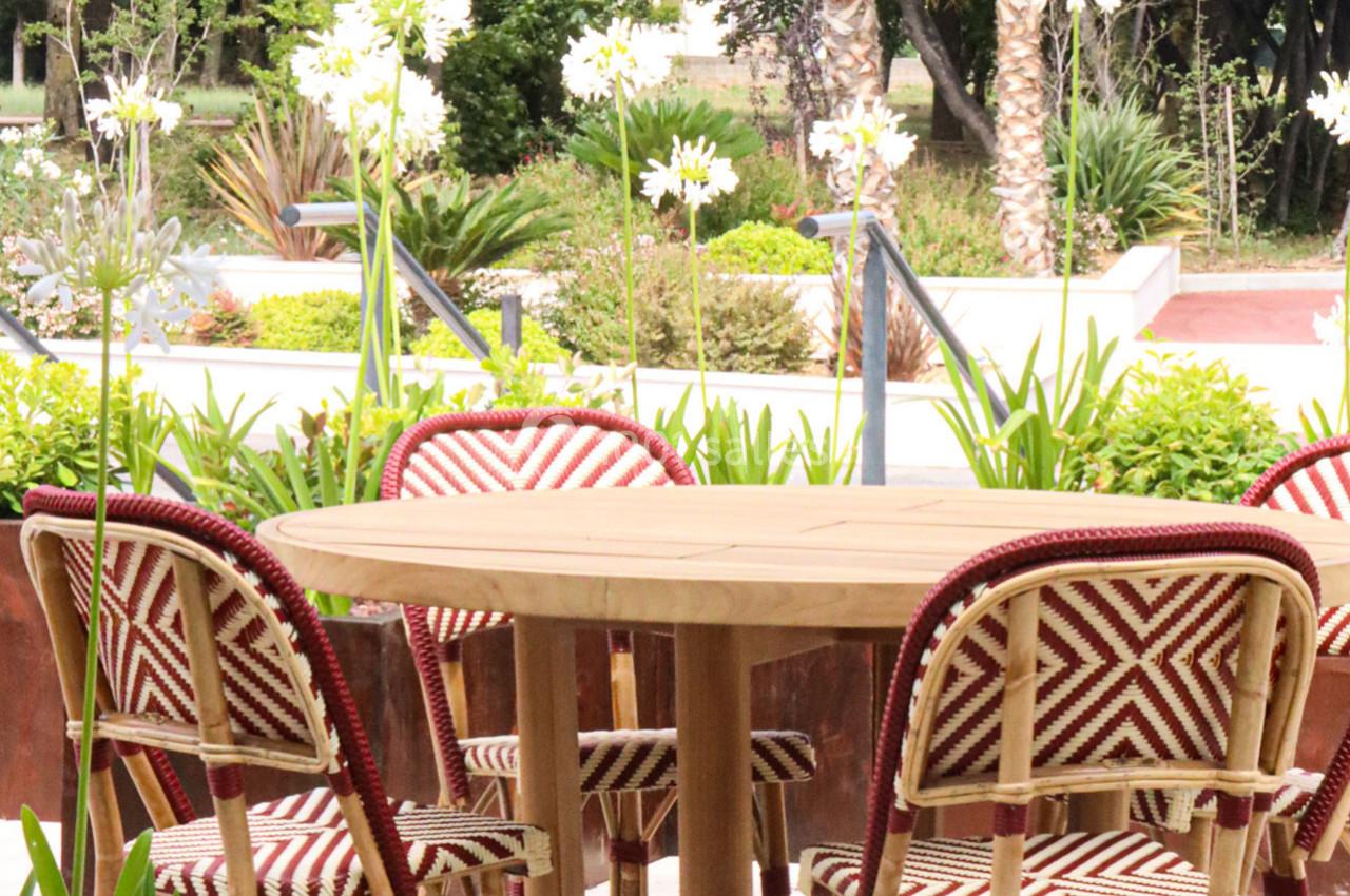 Table ronde en bois entourée de chaises à motifs rouges et blancs, située dans un jardin avec des plantes et des fleurs.
