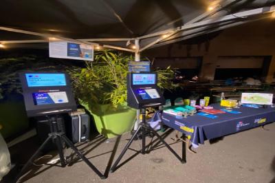 Stand d'information nocturne avec bornes interactives, table d'exposition et éclairage sous une tente.