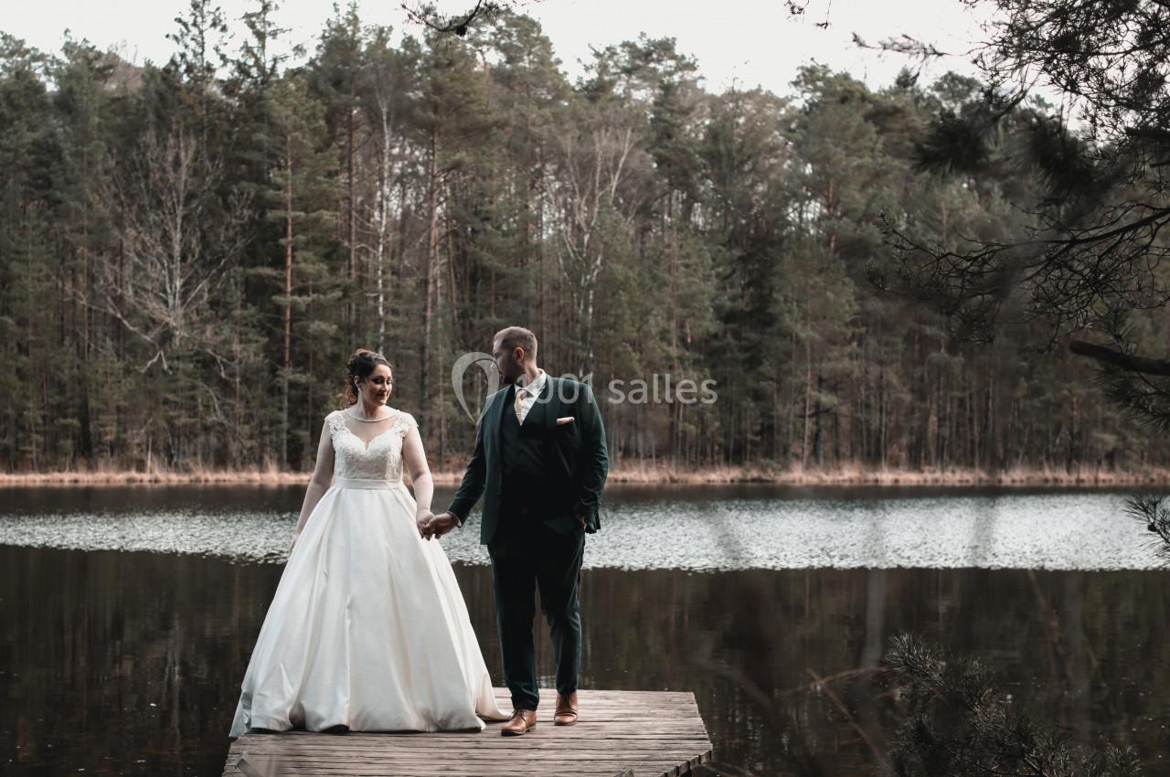 Un couple en tenue de mariage se tient sur un ponton en bois devant un lac entouré d'arbres.