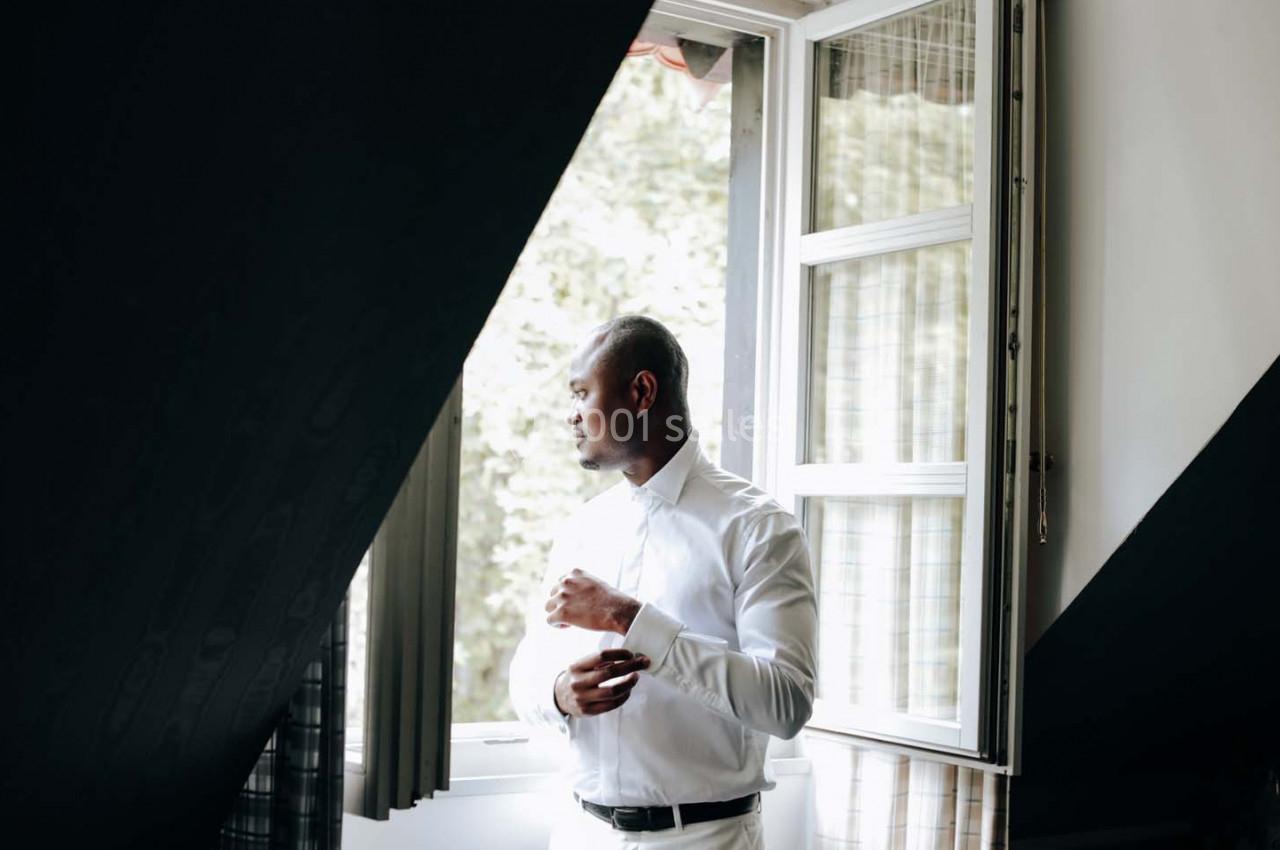 Homme debout près d'une fenêtre ouverte, ajustant sa chemise blanche, avec un fond de verdure à l'extérieur.