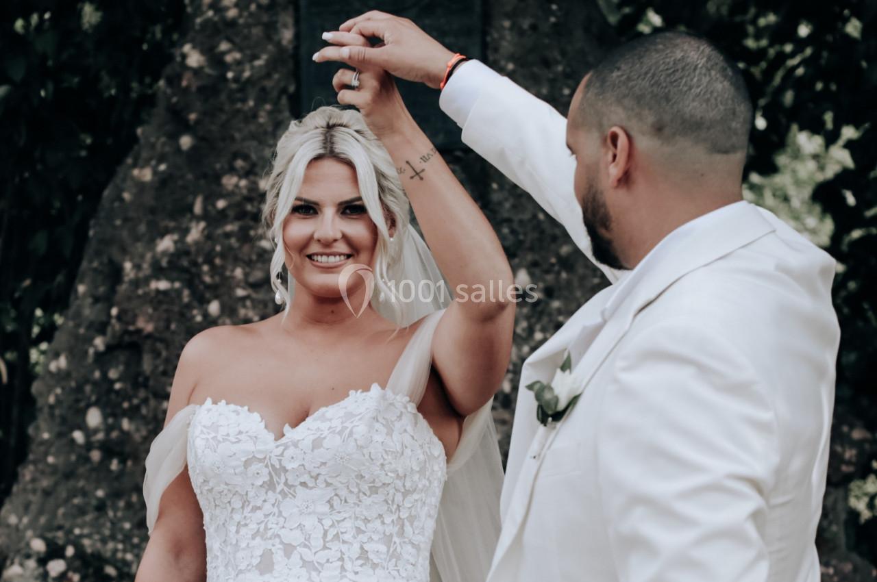 Un couple en tenue de mariage danse devant un arbre, la mariée souriante et le marié en costume blanc.