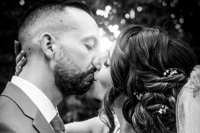 Un couple en tenue de mariage pose devant une voiture ancienne noire et un bâtiment historique entouré de verdure.