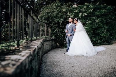 Un couple en tenue de mariage pose devant une voiture ancienne noire et un bâtiment historique entouré de verdure.