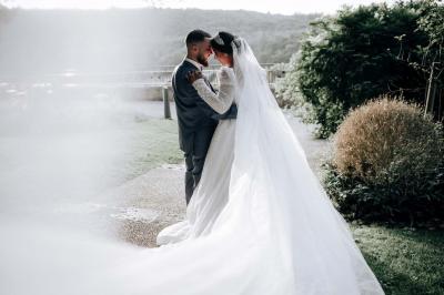 Un couple en tenue de mariage pose devant une voiture ancienne noire et un bâtiment historique entouré de verdure.