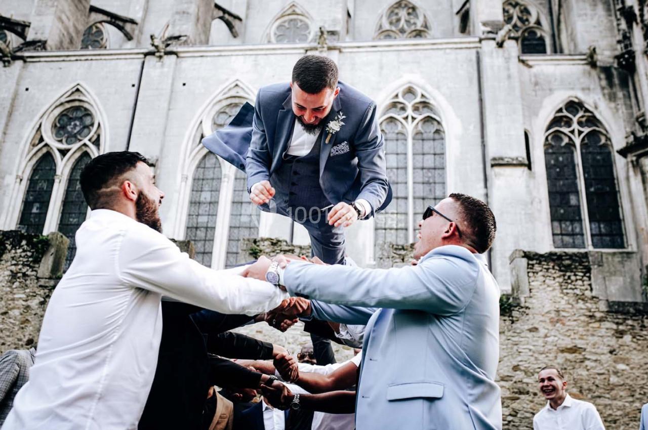 Un homme en costume est lancé en l'air par un groupe de personnes devant une église en pierre.