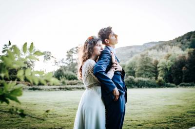 Un couple en tenue de mariage pose devant une voiture ancienne noire et un bâtiment historique entouré de verdure.