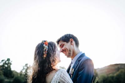 Un couple en tenue de mariage pose devant une voiture ancienne noire et un bâtiment historique entouré de verdure.