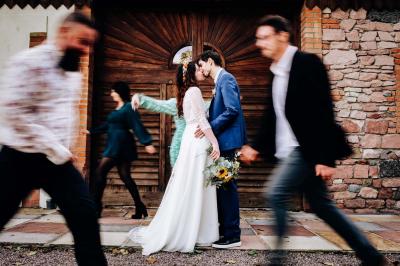 Un couple en tenue de mariage pose devant une voiture ancienne noire et un bâtiment historique entouré de verdure.