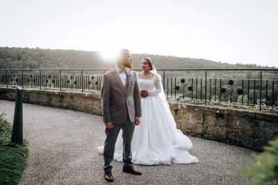 Un couple en tenue de mariage pose devant une voiture ancienne noire et un bâtiment historique entouré de verdure.