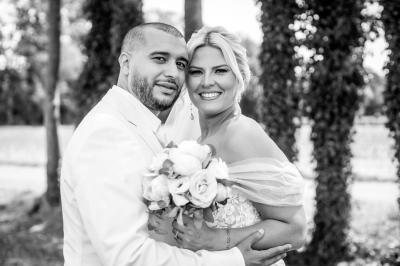 Un couple en tenue de mariage pose devant une voiture ancienne noire et un bâtiment historique entouré de verdure.