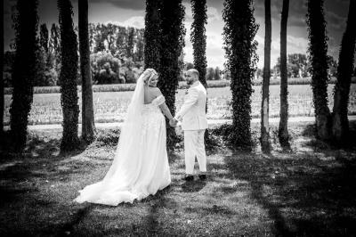 Un couple en tenue de mariage pose devant une voiture ancienne noire et un bâtiment historique entouré de verdure.