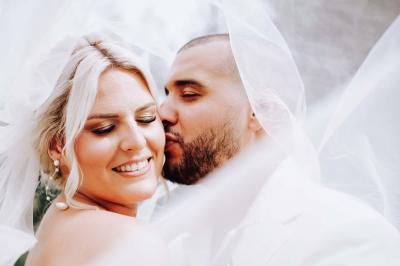 Un couple en tenue de mariage pose devant une voiture ancienne noire et un bâtiment historique entouré de verdure.