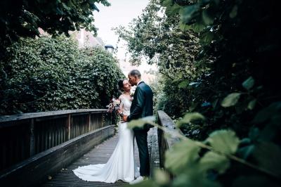 Un couple en tenue de mariage pose devant une voiture ancienne noire et un bâtiment historique entouré de verdure.