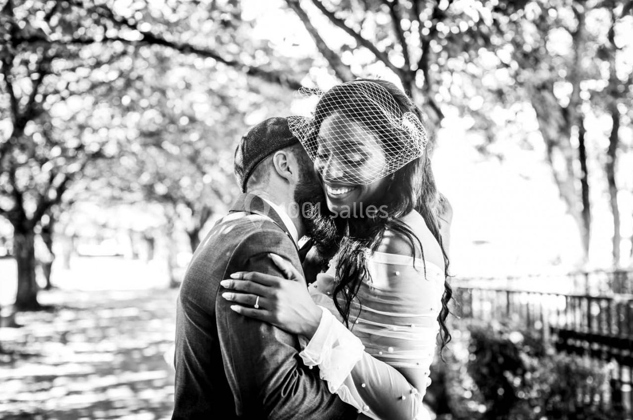 Un couple souriant s'enlace sous des arbres dans une allée ensoleillée, en noir et blanc.