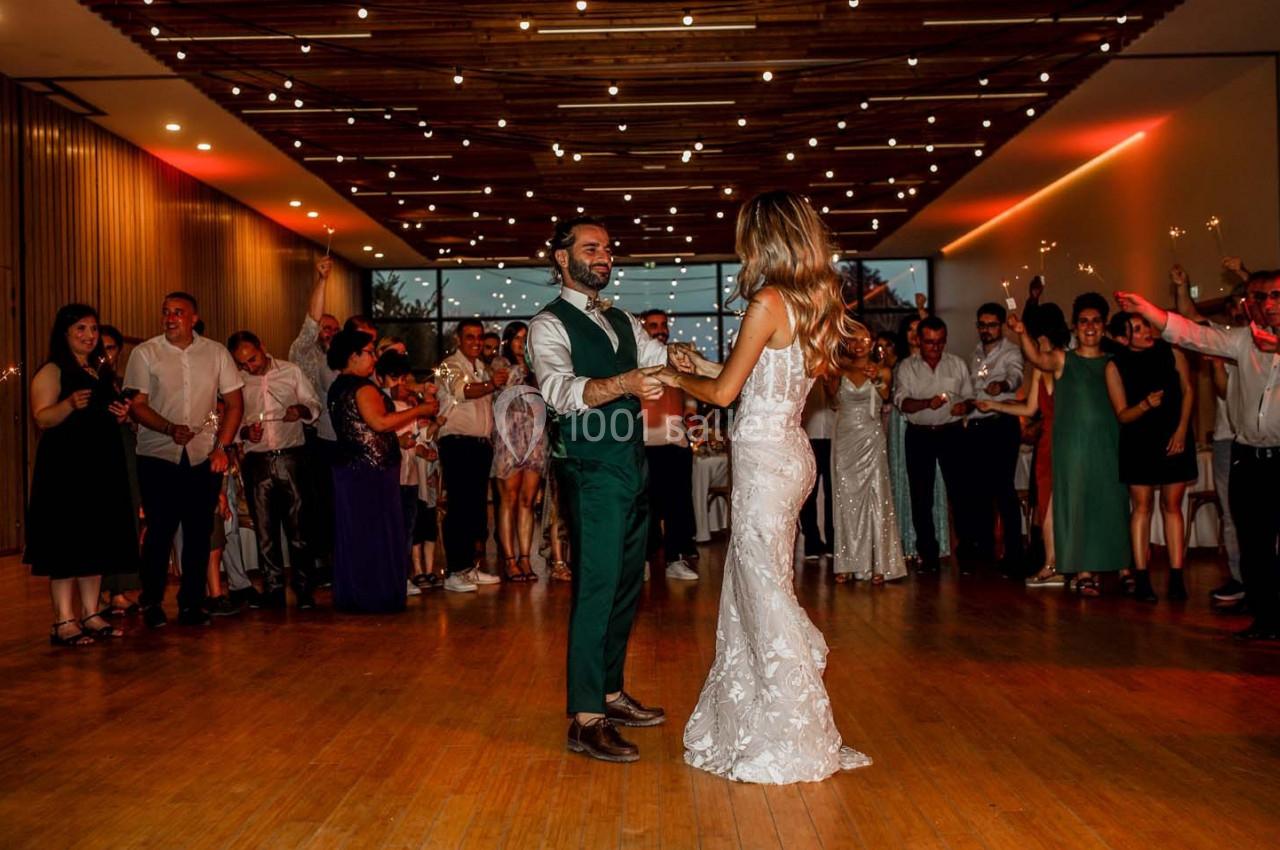Un couple danse au centre d'une salle éclairée, entouré d'invités applaudissant.