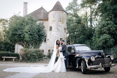 Un couple en tenue de mariage pose devant une voiture ancienne noire et un bâtiment historique entouré de verdure.