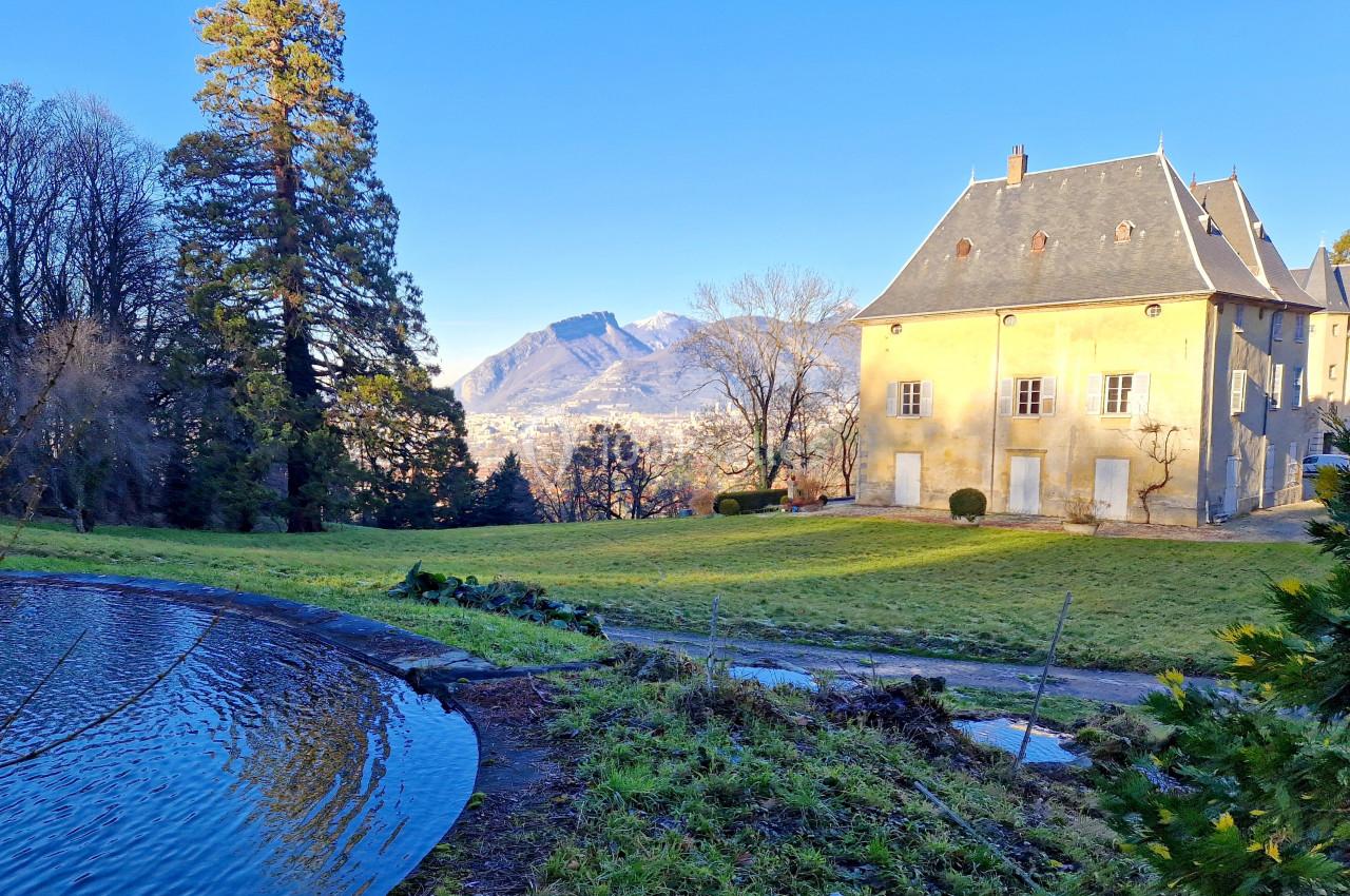 Vue d'un bâtiment ancien entouré de verdure, avec un étang au premier plan et des montagnes en arrière-plan.