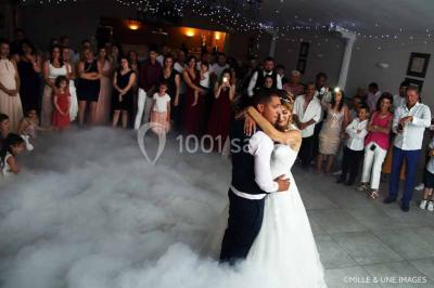 Un couple de mariés danse au centre d'une salle entourée d'invités, avec un sol recouvert de fumée légère.