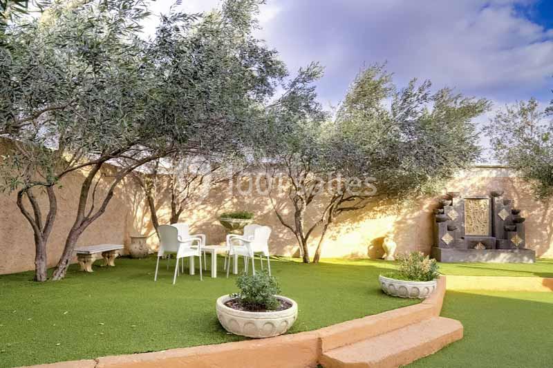 Jardin avec pelouse artificielle, oliviers, mobilier de jardin blanc et fontaine murale décorative en pierre.