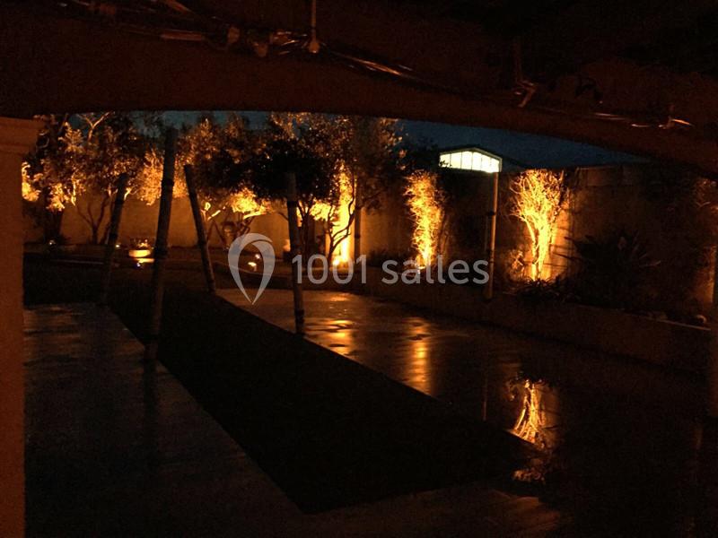 Cour extérieure illuminée la nuit, avec des arbres et des murs éclairés par des lumières chaudes.