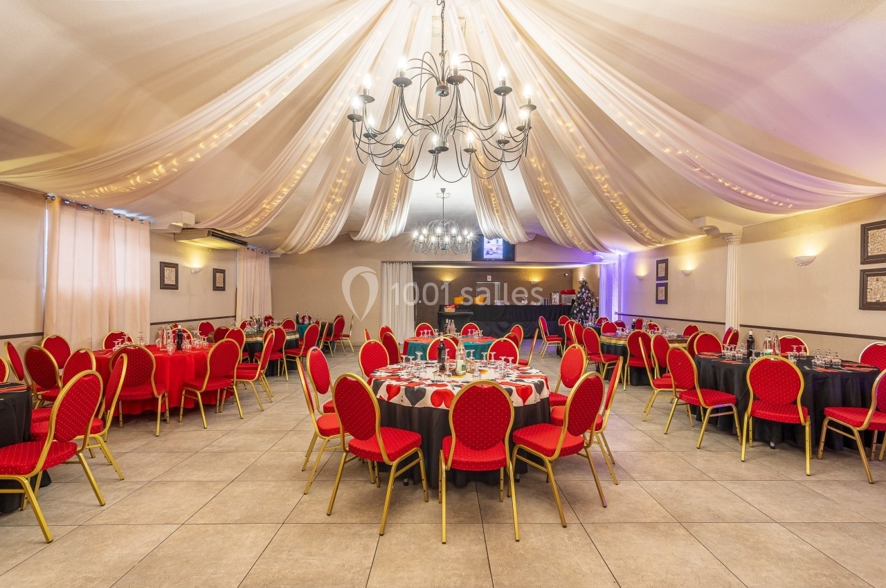 Salle de réception décorée avec des tables rondes dressées, chaises rouges et plafond orné de drapés et lustres.