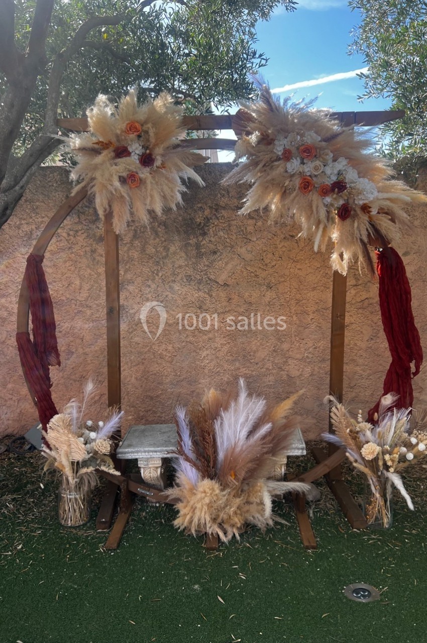 Arche décorative en bois ornée de fleurs séchées et pampas, installée sur une pelouse devant un mur en pierre.
