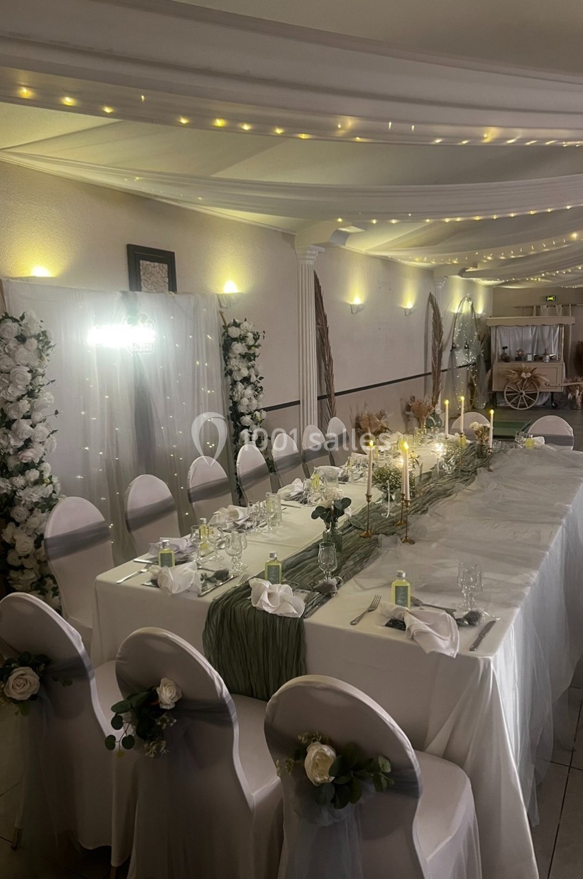 Salle de réception décorée avec une table de banquet ornée de bougies, fleurs blanches et guirlandes lumineuses au plafond.