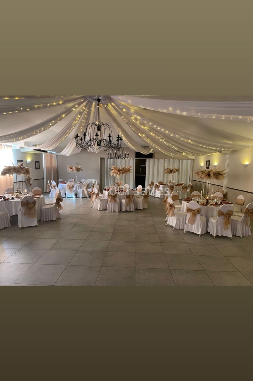 Salle de réception décorée avec des tables rondes, nappes blanches, chaises ornées de nœuds beige et guirlandes lumineuses…