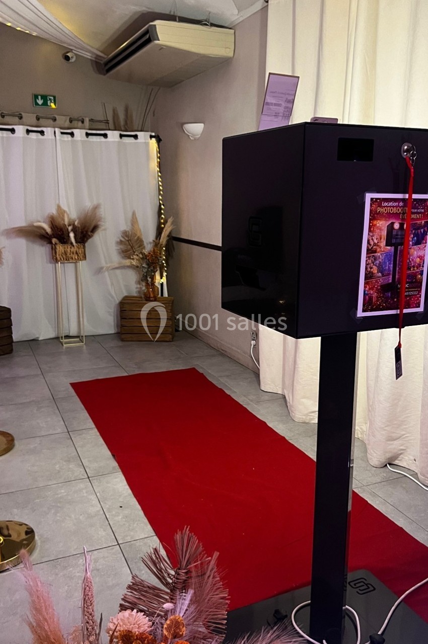 Borne photo noire sur pied devant un tapis rouge, décorée de fleurs séchées dans une salle lumineuse.