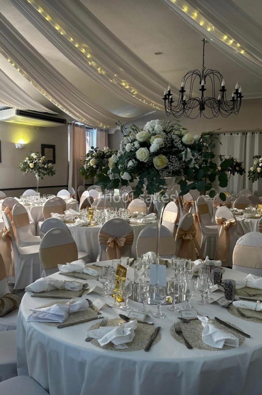 Salle de réception décorée avec des tables rondes, nappes blanches, chaises ornées de nœuds beige et centres de table…