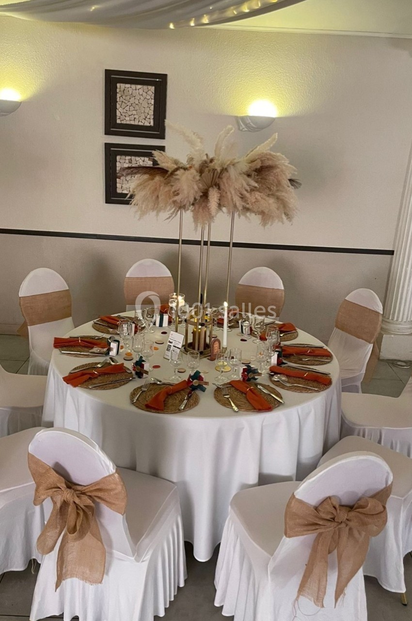 Table ronde décorée pour un événement, avec nappes blanches, chaises ornées de nœuds en toile de jute et centre de table…