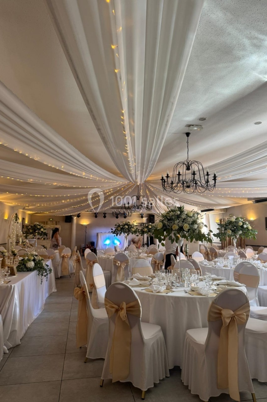 Salle de réception décorée avec des tables rondes, nappes blanches, chaises ornées de rubans dorés et guirlandes lumineuses.