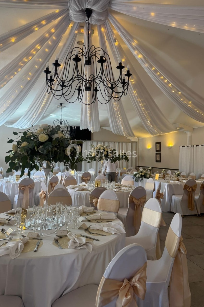 Salle de réception décorée avec des tables rondes, nappes blanches, chaises ornées de rubans dorés et guirlandes lumineuses…
