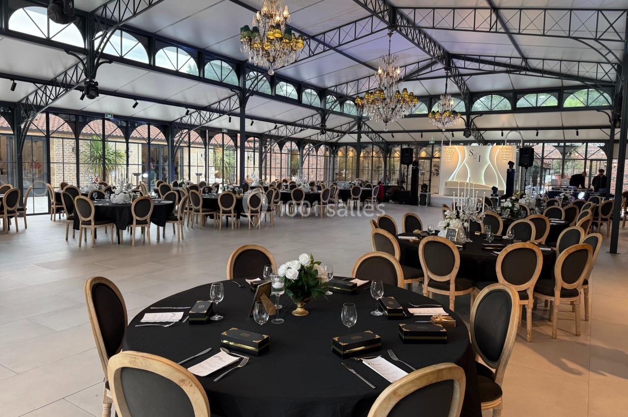 Salle de réception élégante avec tables rondes dressées, chaises en bois, nappes noires et lustres suspendus.
