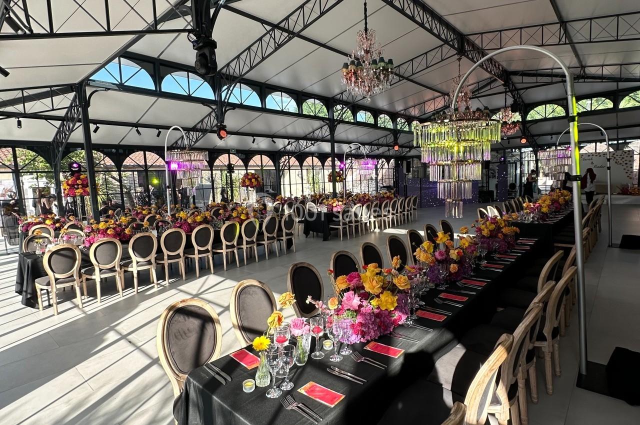 Grande salle lumineuse avec tables décorées de nappes noires, fleurs colorées et chaises élégantes sous une structure…