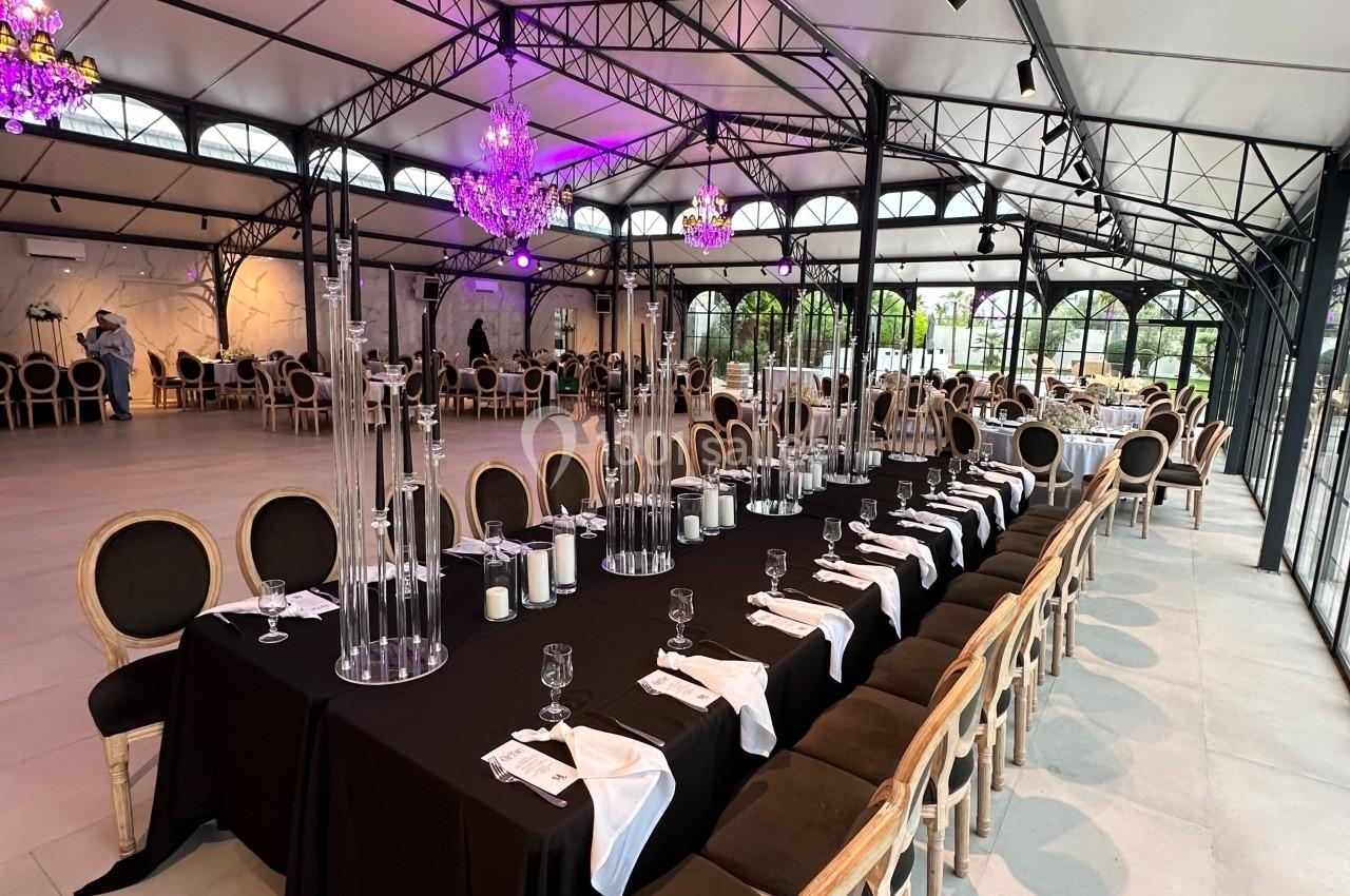 Grande table rectangulaire dressée avec une nappe noire, des chandeliers et des chaises élégantes dans une salle lumineuse.