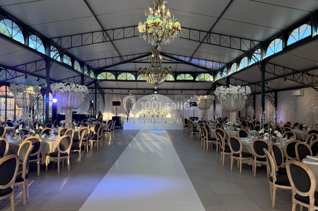 Salle de réception élégante avec tables décorées, chandeliers suspendus et allée centrale menant à une scène ornée de fleurs.