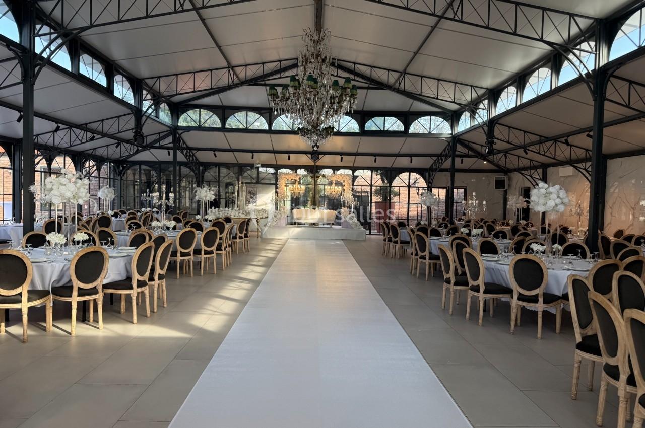Salle de réception élégante avec tables rondes dressées, chaises noires et dorées, et un lustre central imposant.